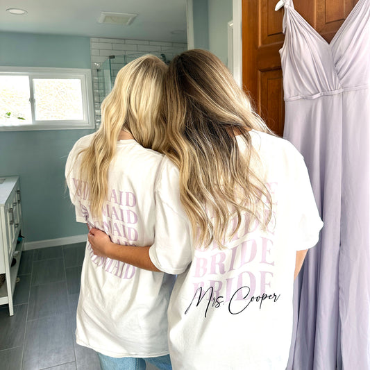 a woman and her bridesmaid both wearing white comfort colors t-shirts with a retro wave printed bridesmaid and bride design on the back. the bride's shirt featuring her married name along the bottom