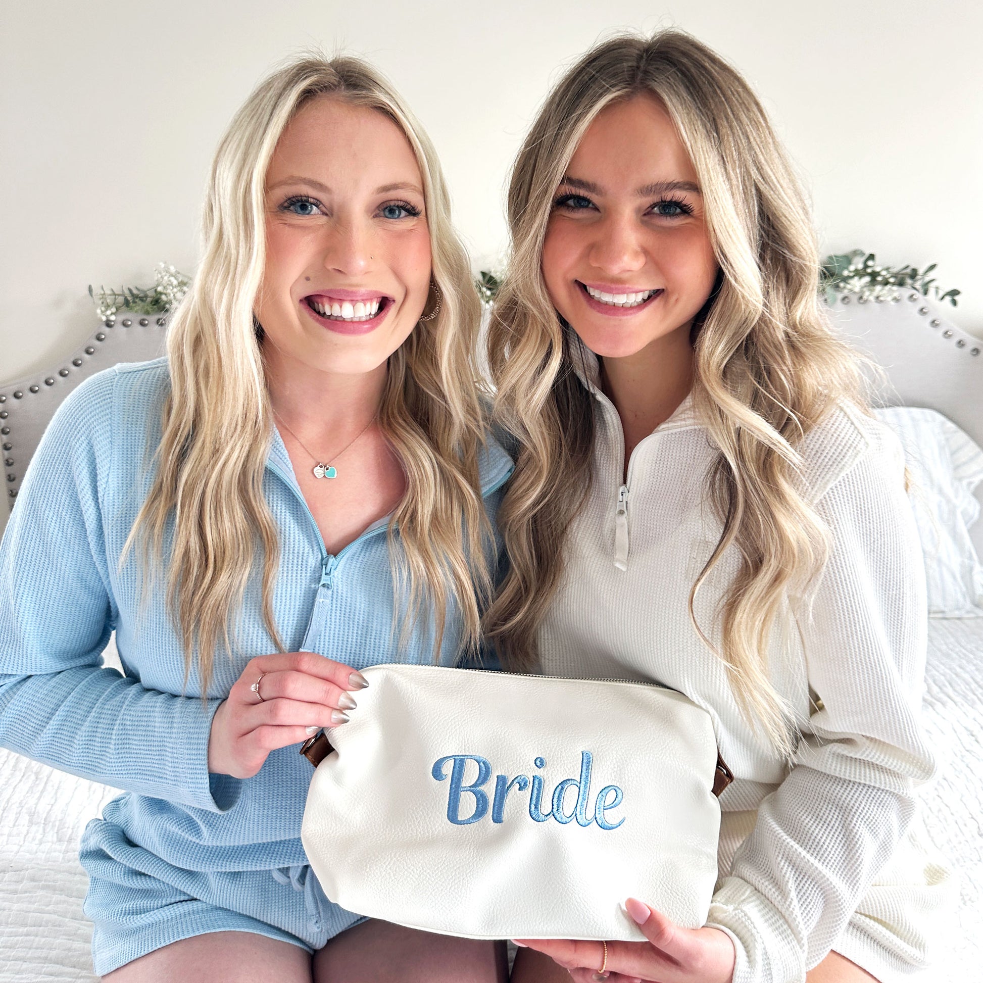 bride and bridesmaid holding a white addison dopp kit with embroidered bride in baby blue thread on the center of the bag