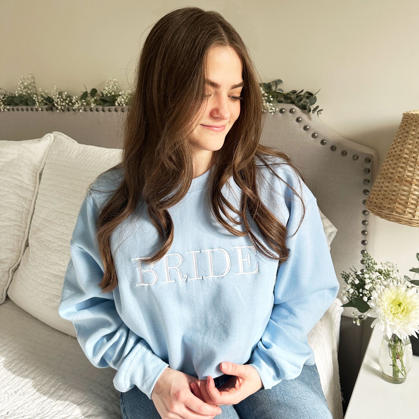Young woman wearing a light blue crewneck sweatshirt with all caps bride embroidered in white thread