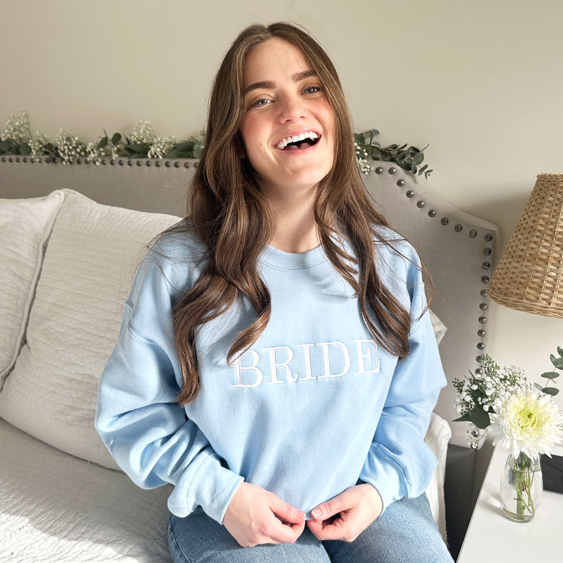 Young woman wearing a light blue crewneck sweatshirt with bride embroidered in all caps and white thread