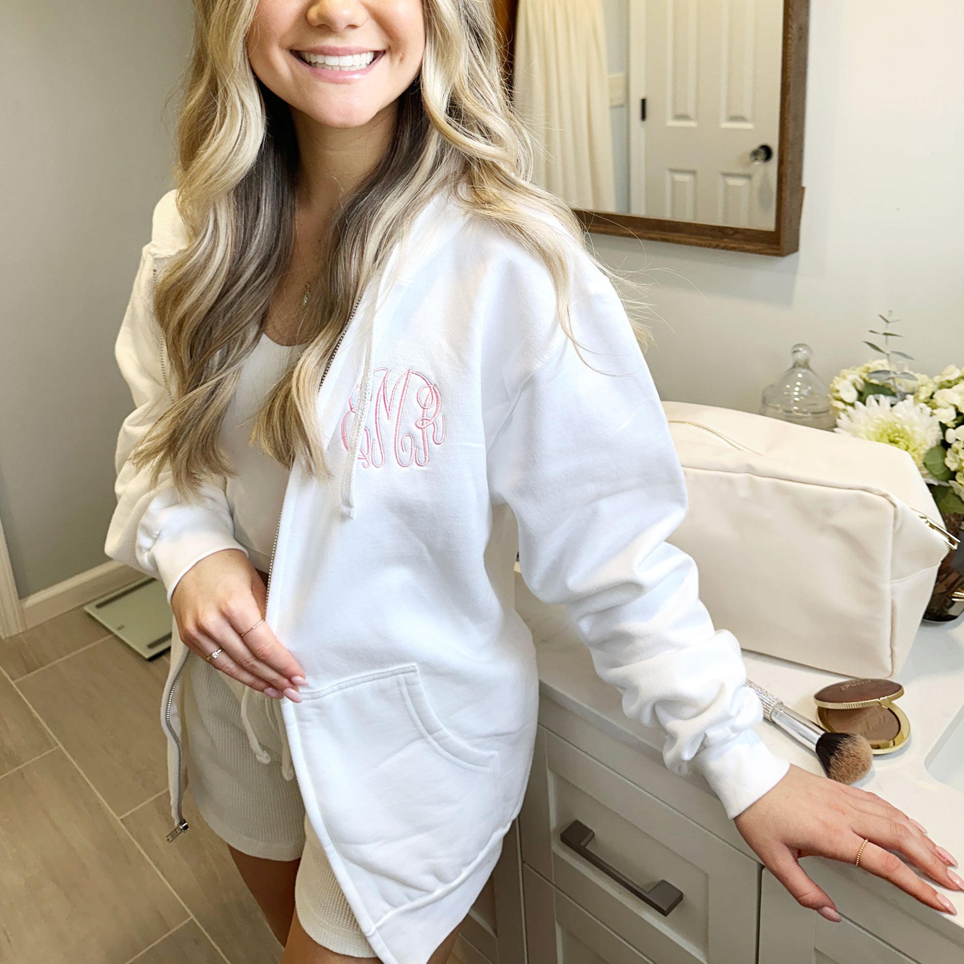 young woman wearing a white full zip hooded sweatshirt with monogram embroidery on the left chest in baby pink thread