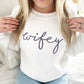 Close up of a woman wearing a white crewneck sweatshirt with script wifey embroidered in smoky orchid thread