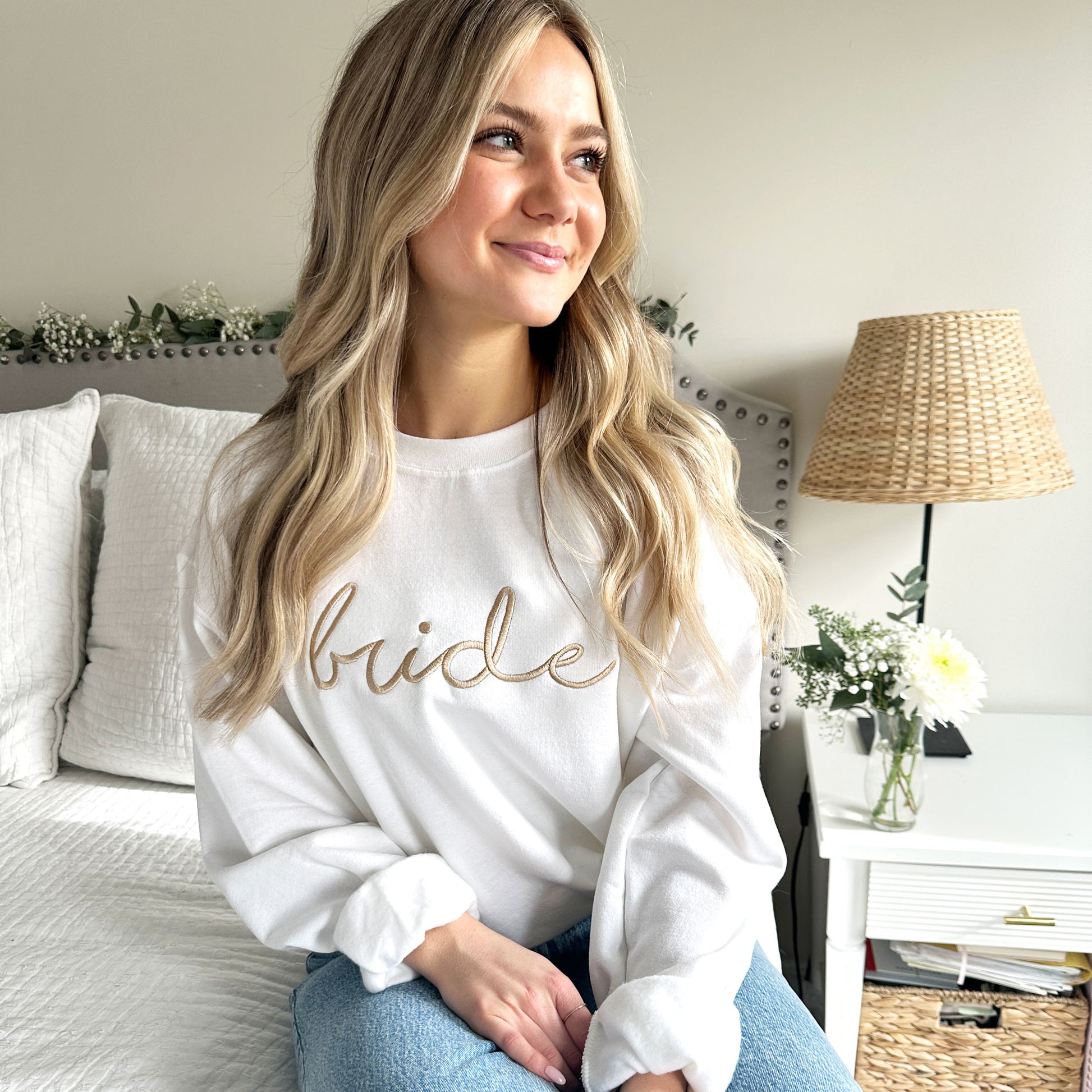 Young woman modeling a white crewneck sweatshirt with bride embroidered across the chest in camel thread