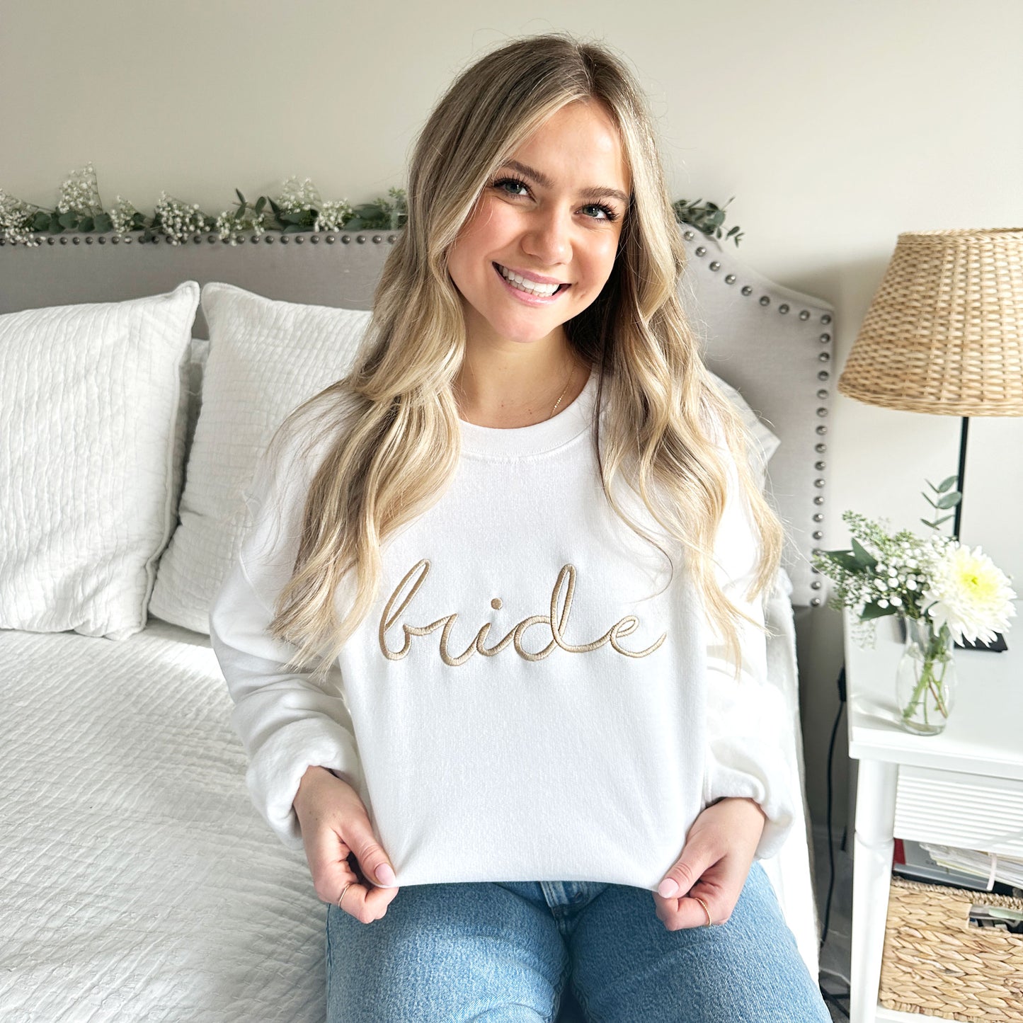 Young woman wearing a white crewneck sweatshirt that features bride embroidered in script font across the chest in camel thread