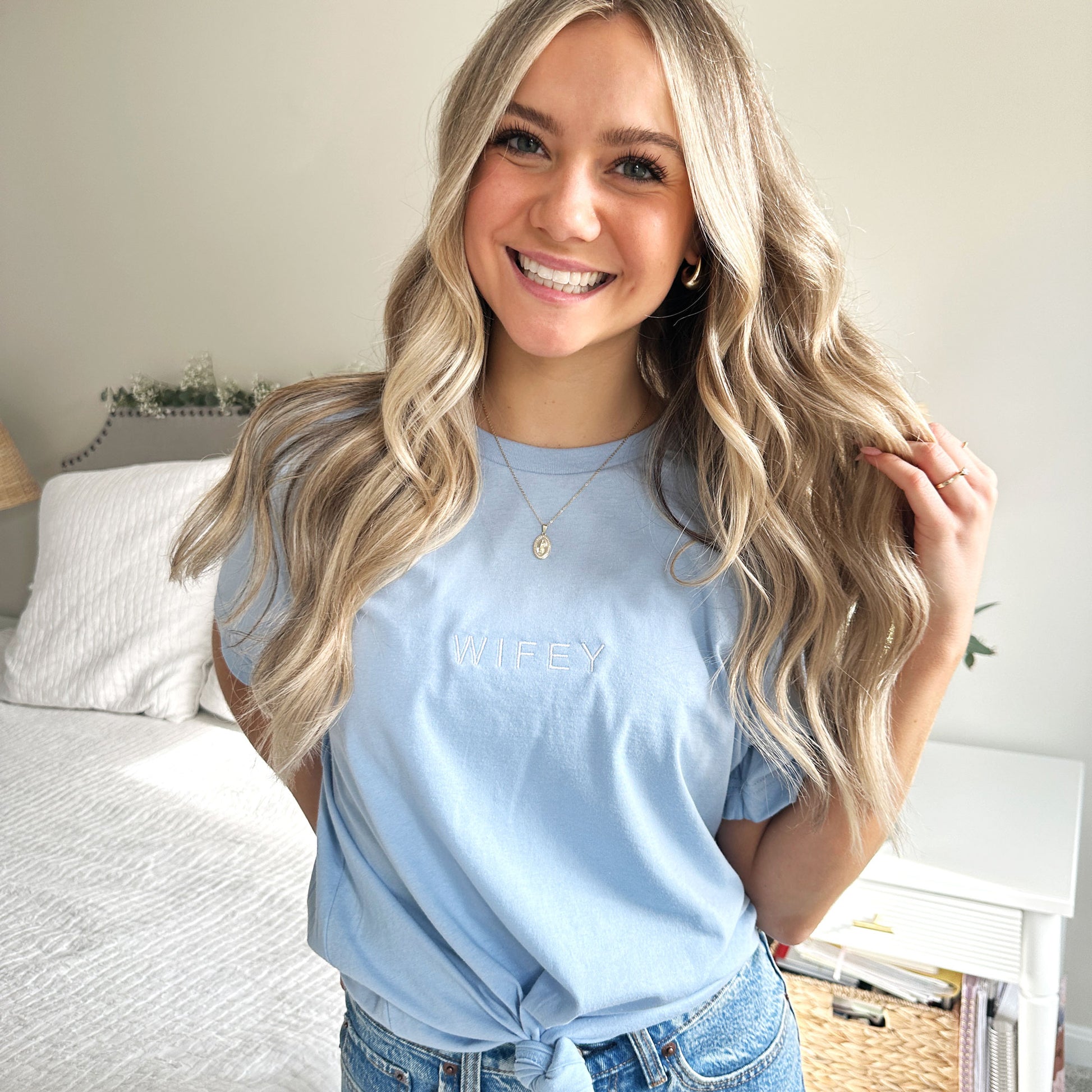 young woman wearing a baby blue crewneck bella and canvas tshirt with wifey embroidered in all caps in white thread small across the chest