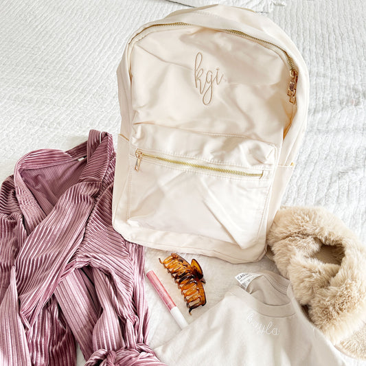 creme backpack on a bed with a robe, t-shirt, claw clip, lip gloss, and slippers