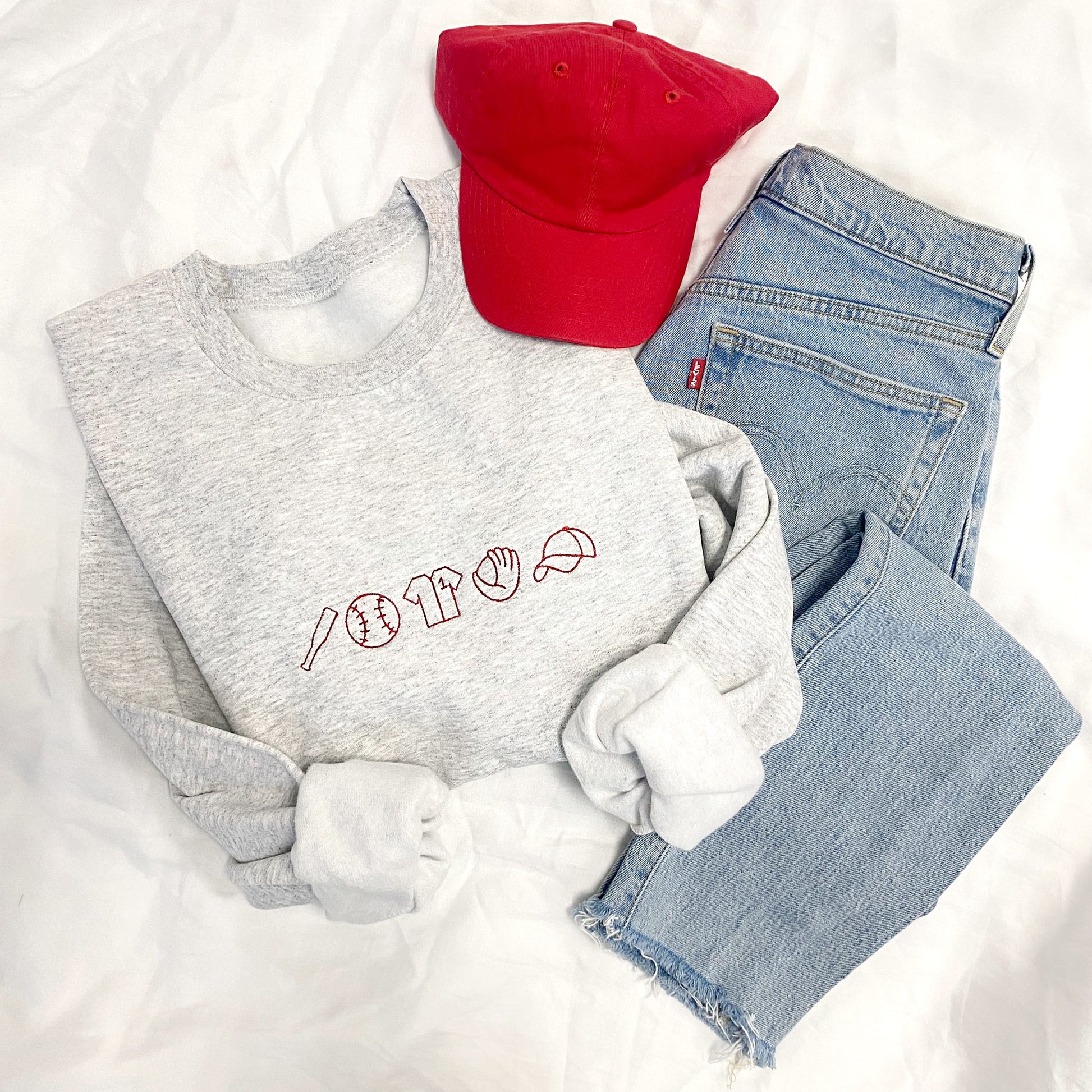 Ash crewneck sweatshirt with red embroidered baseball icons styled with a red baseball cap and blue jeans
