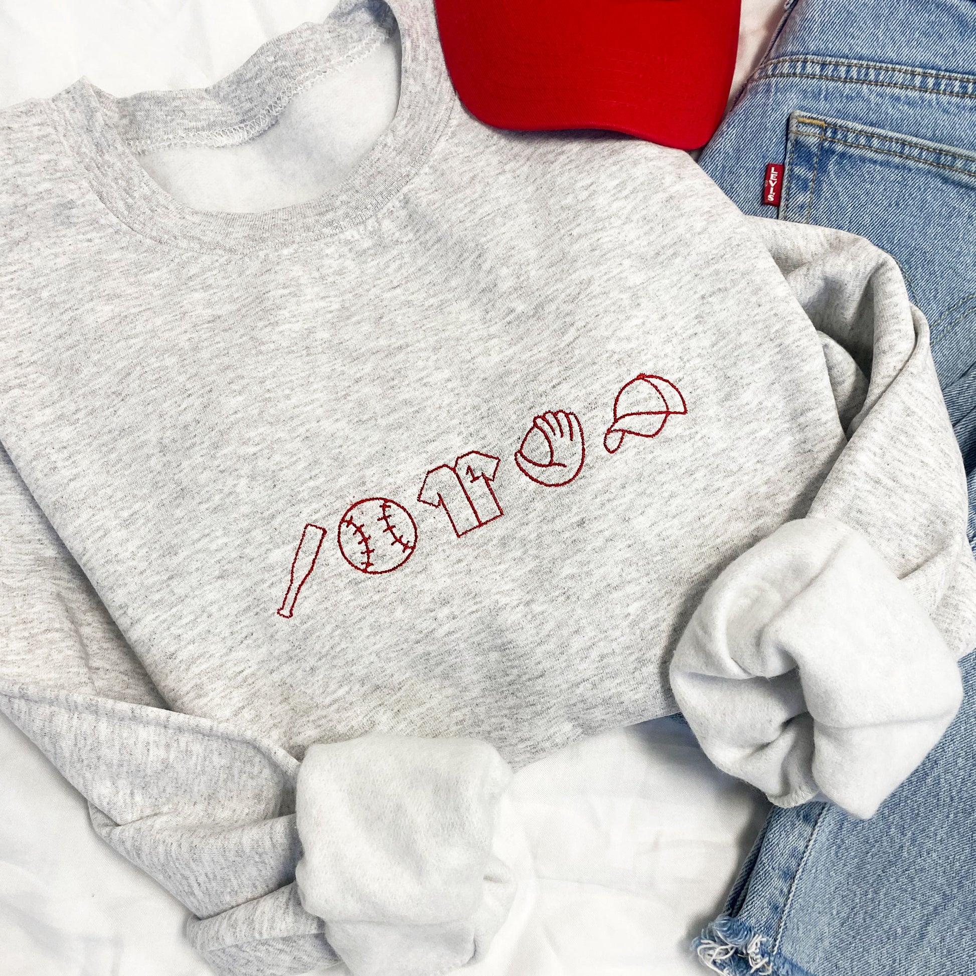 Close op of styled flat lay of an ash crewneck sweatshirt with embroidered baseball icons in red thread
