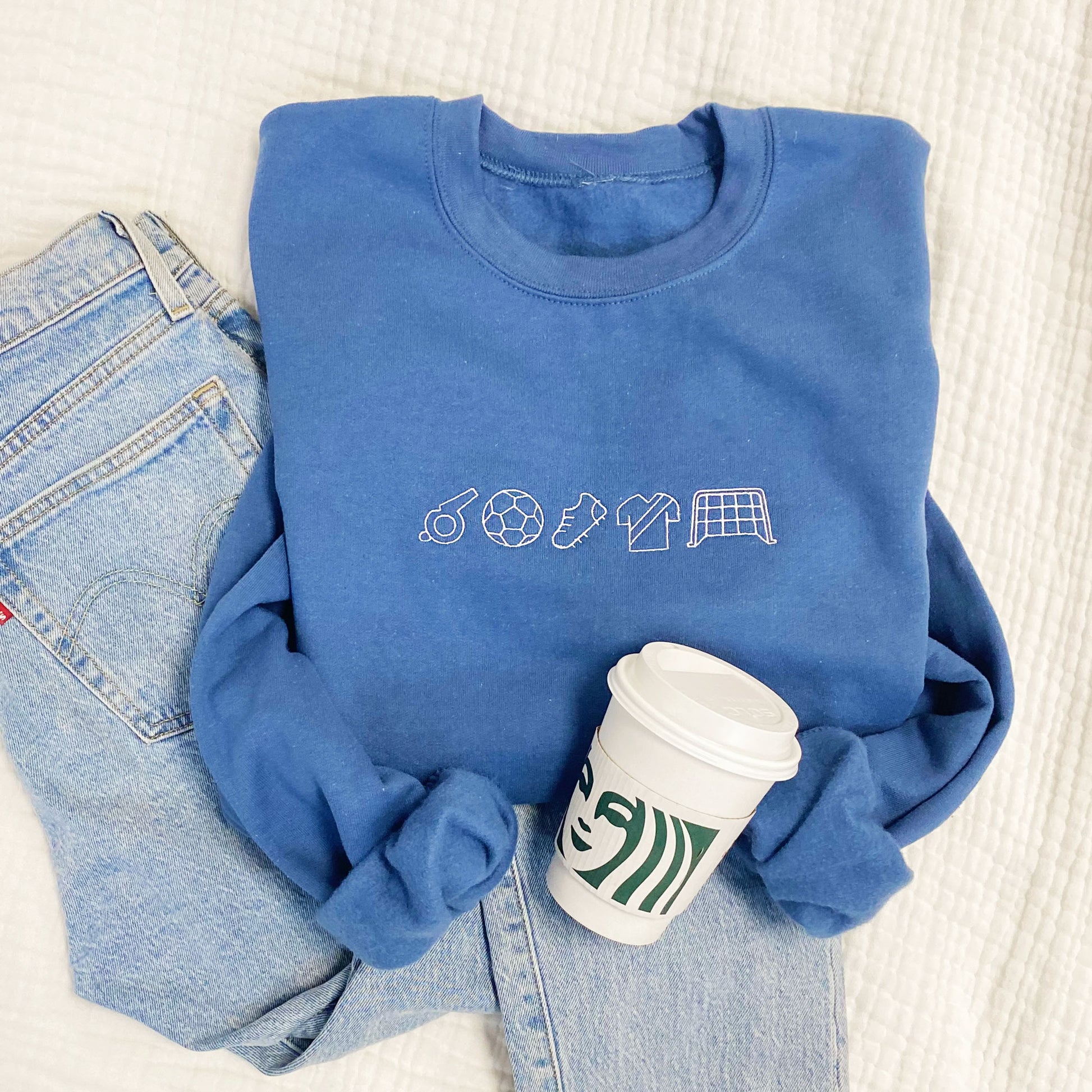 styled flat lay of indigo sweatshirt with jeans and a starbucks cup. the sweatshirt features white soccer icons embroidered across the chest