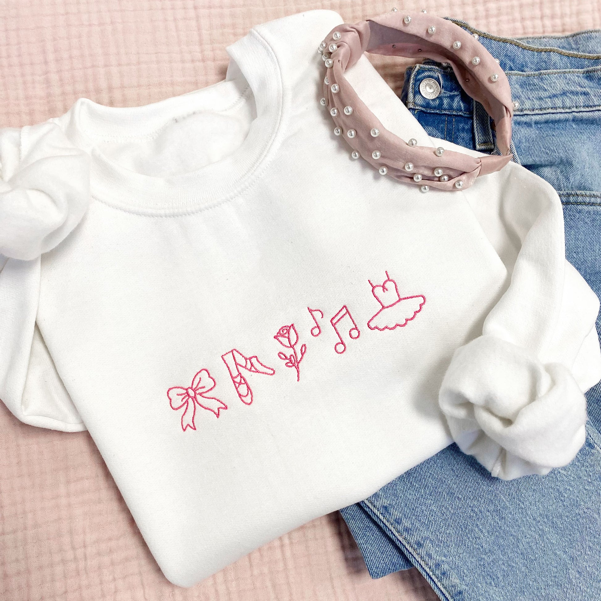 Styled flat lay with a white crewneck sweatshirt with pink embroidered dance icons across the center chest