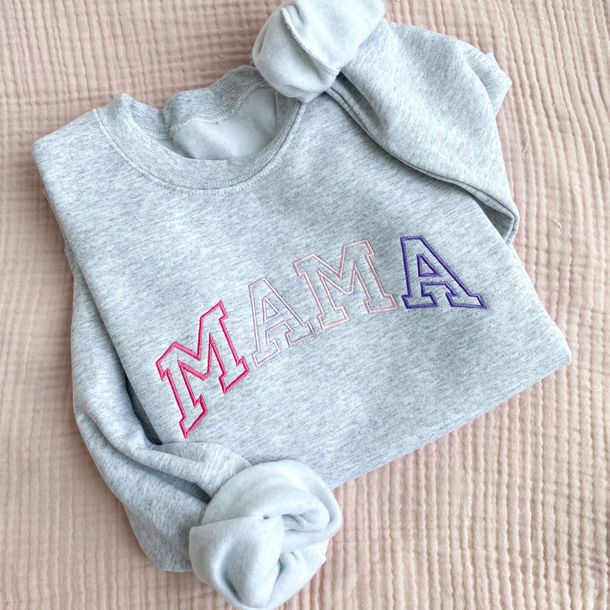 flat lay of an ash crewneck sweatshirt with embroidered athletic block mama in alternating shades of pink and purple