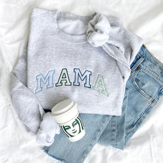 flat lay photo of an ash crewneck sweatshirt with large embroidered athletic block MAMA in alternating blues and greens styled with jeans and a Starbucks cup