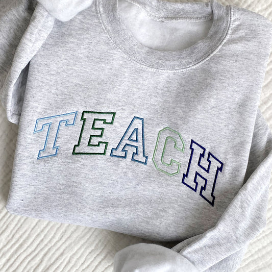 flat lay photo of an ash crewneck sweatshirt with athletic block outline embroidered TEACH in blues and greens