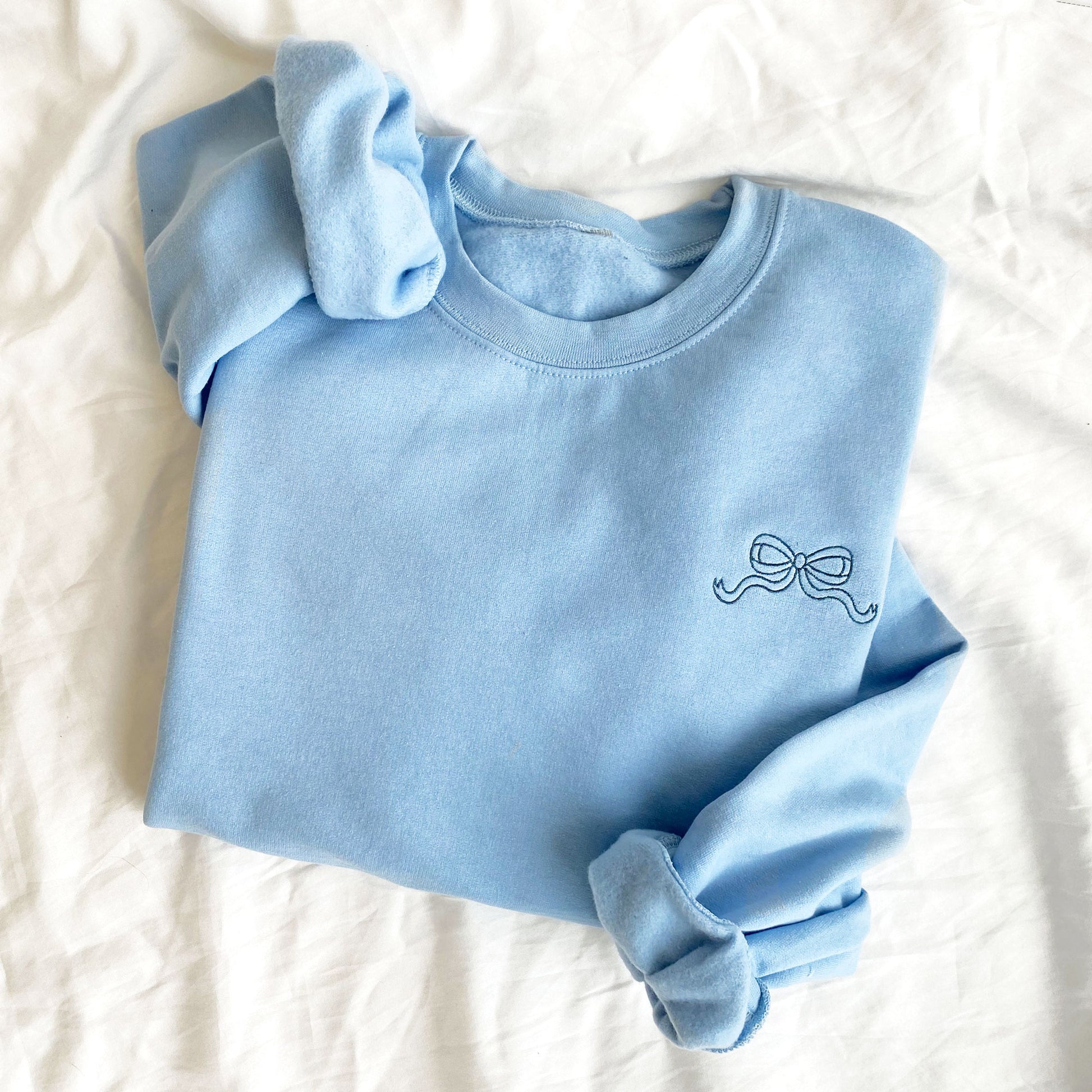 flat lay of light blue crewneck sweatshirt with mini outlined bow in blue on the left chest