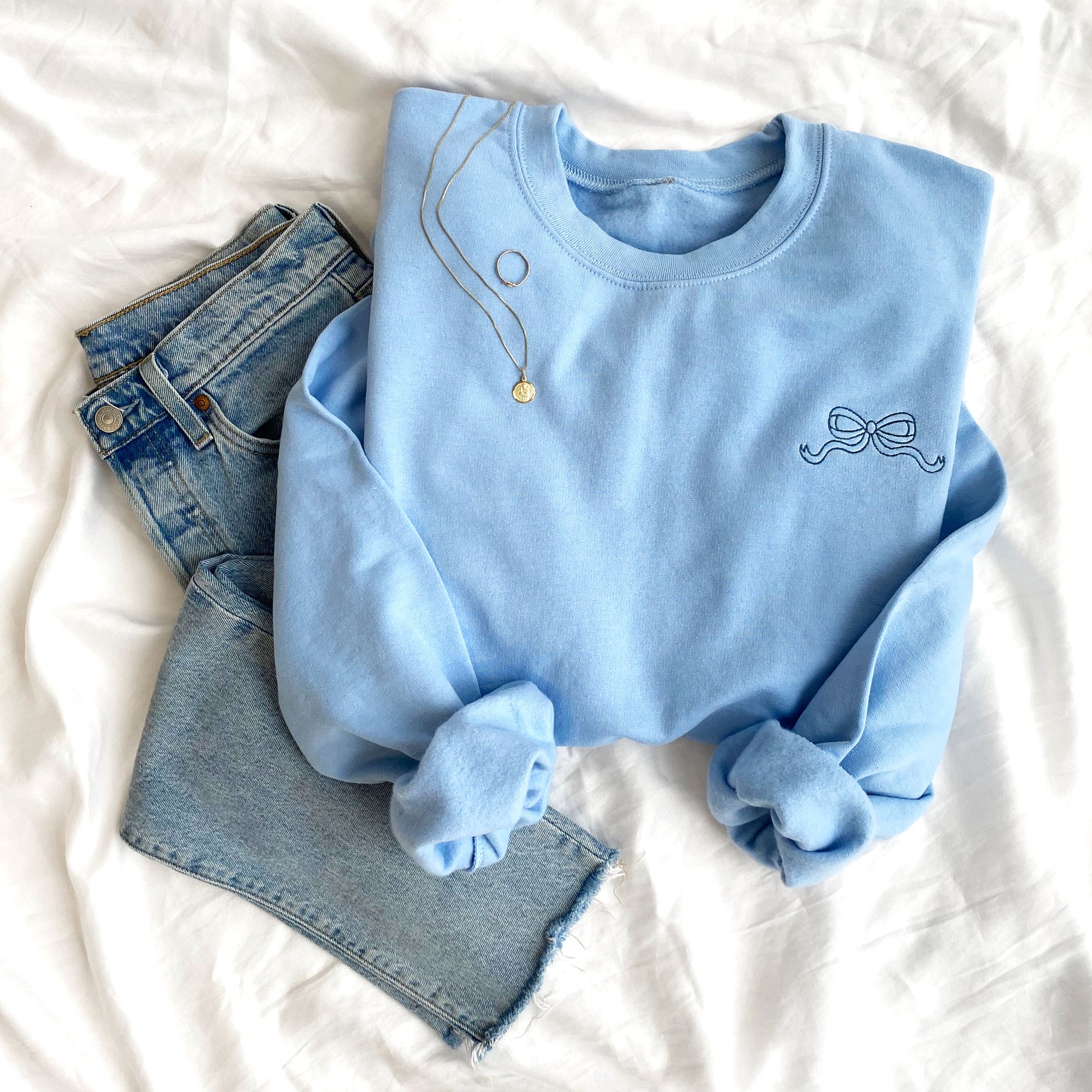 styled flat lay of jeans gold jewelry and a light blue crewneck sweatshirt with left chest embroidery of an outlined blue bow