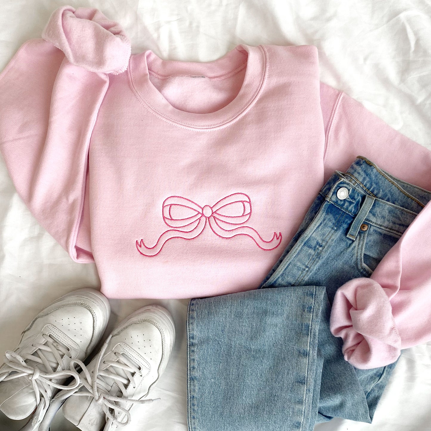 styled flat lay of a light pink crewneck sweatshirt with pink outline embroidered bow. Styled with jeans and white sneakers