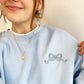young woman wearing a light blue crewneck sweatshirt with mini outlined bow on the left chest embroidered in french blue thread