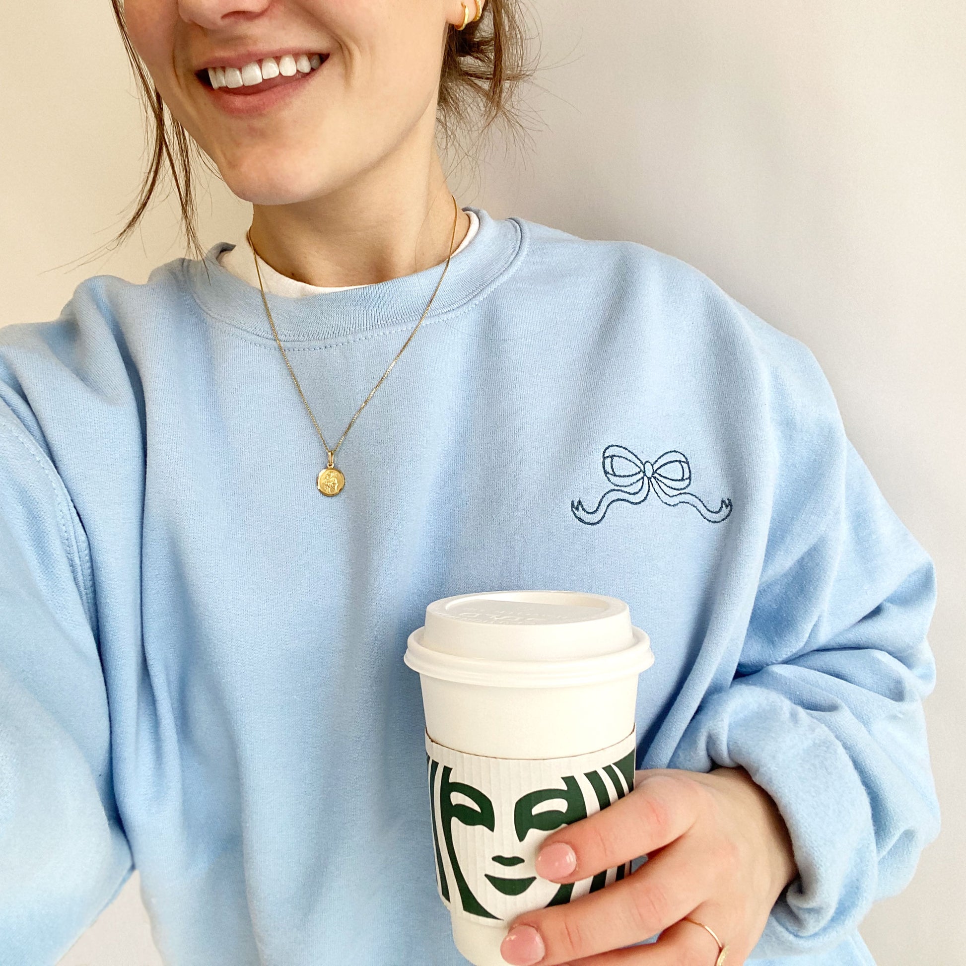 young woman wearing a light blue crewneck sweatshirt with mini outlined embroidered bow in french blue thread on the left chest