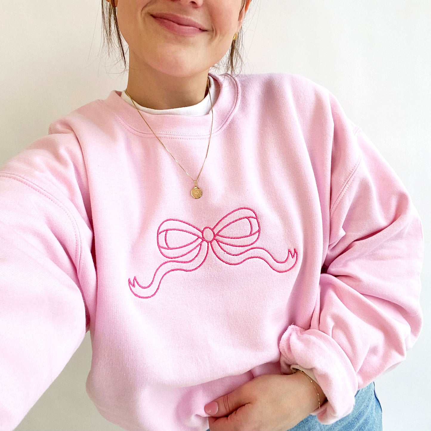 Woman wearing a light pink crewneck sweatshirt with large outline bow embroidered across the chest in pink thread