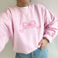 Young woman wearing a light pink crewneck sweatshirt with pink embroidered outline bow across the chest