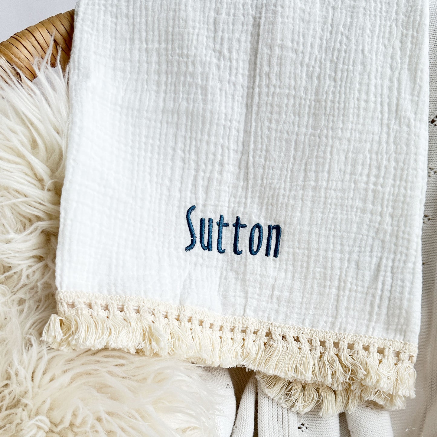 close up of a muslin baby blanket with decorative trim and custom embroidered name design on the corner 
