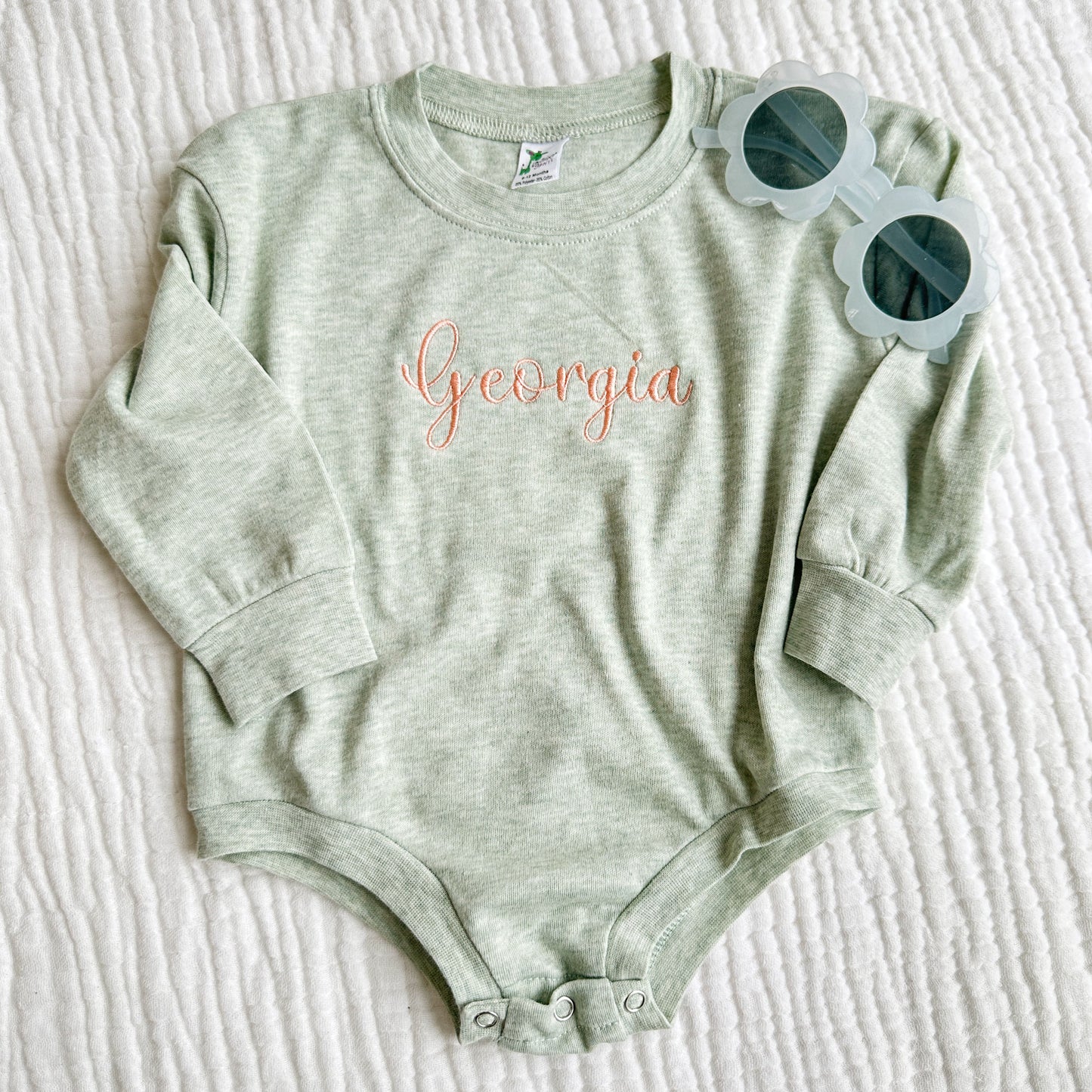 an infant bubble romper in a heather sage with a custom center chest name embroidery in a girly script font and a coral pink thread