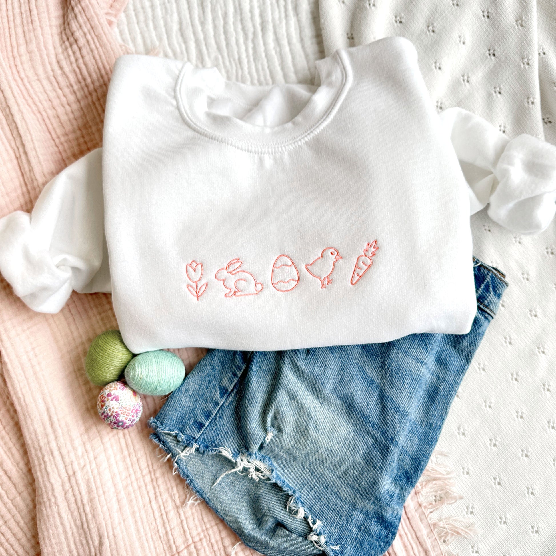 Styled flat lay of a white crewneck sweatshirt with playful easter icons embroidered in coral pink thread across the chest