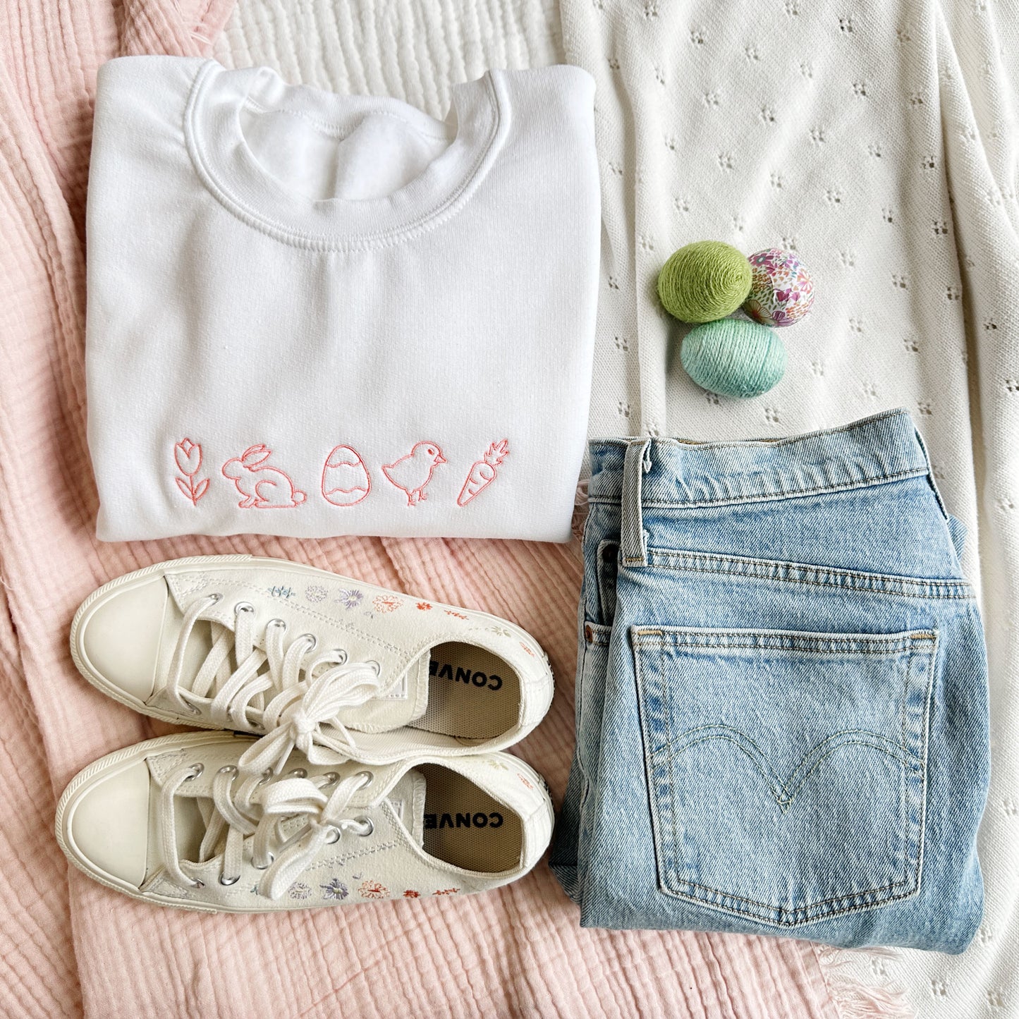 Flat lay of jeans, sneaker, and Easter eggs styled with a white crewneck sweatshirt that has small easter icons embroidered across the chest in coral pink thread