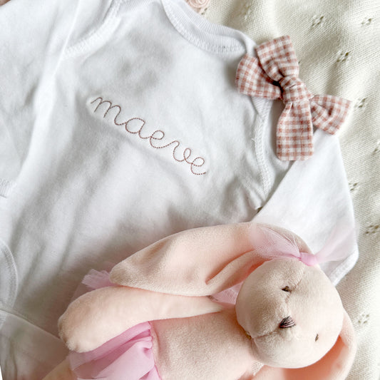 close up of a white baby bodysuit with a personalized name embroidered in a mauve thread and stitched script font across the chest