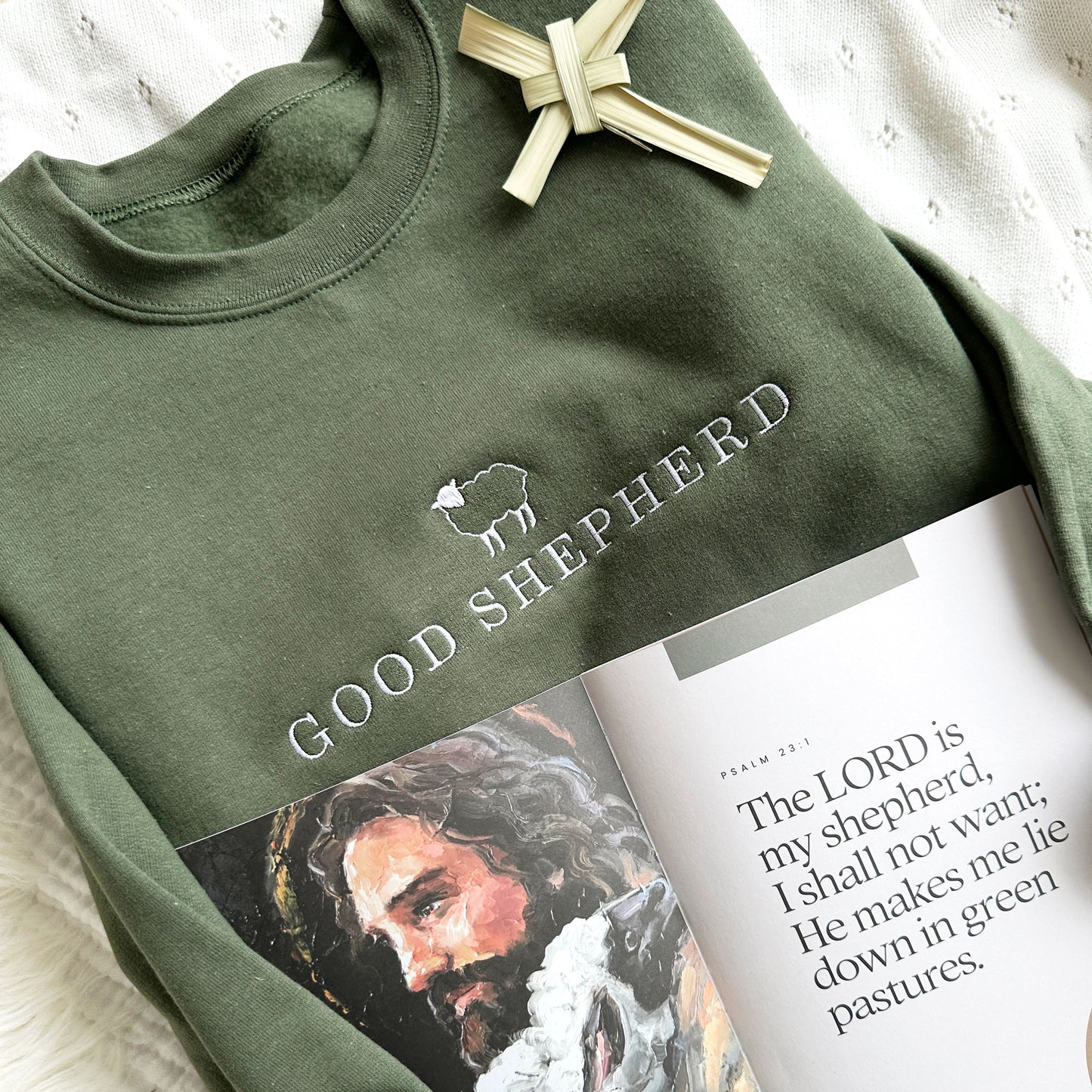 Embroidered sheep and good shepherd across an olive crewneck sweatshirt styled with an open book 