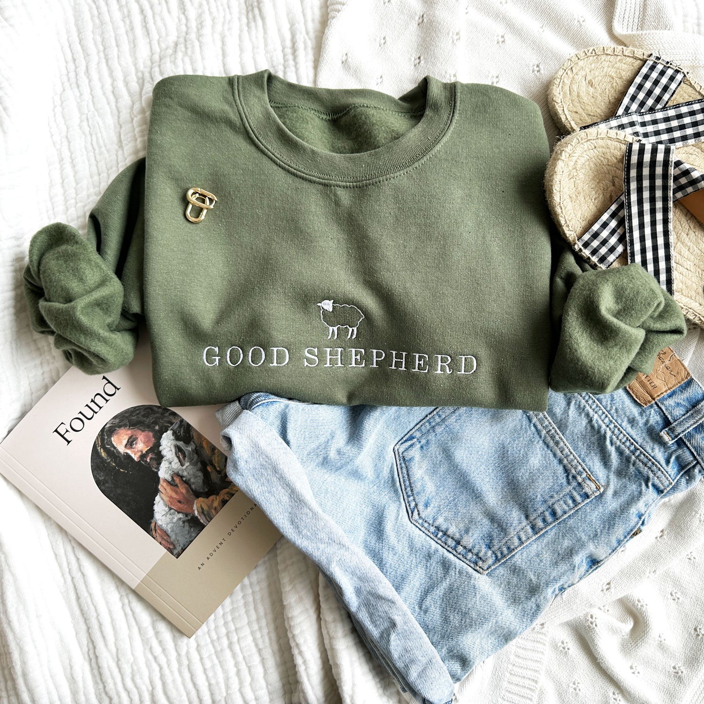 Styled flat lay of jeans, sandals, religious book, and olive crewneck sweatshirt. On the sweatshirt is a little sheep above the words good shepherd embroidered in white thread 