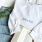 styled flat lay of jeans and a bible with a white crewneck sweatshirt with bible verse John 3:16 embroidered in french blue thread