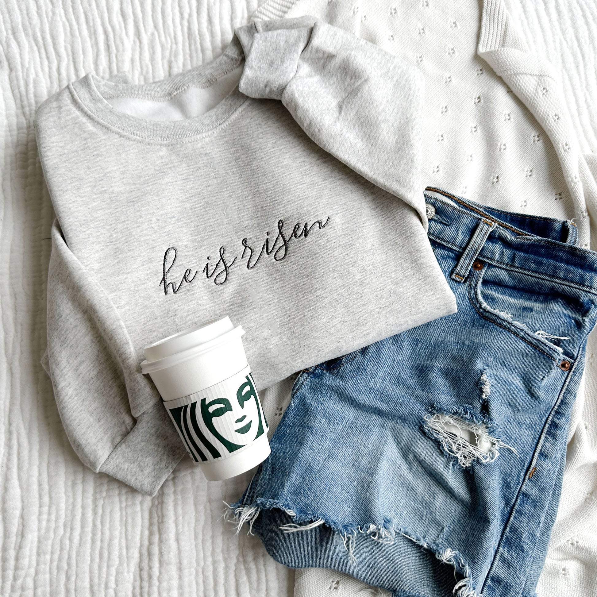 Embroidered Ash crewneck sweatshirt with he is risen embroidered in a script font and gray thread, styled with a Starbucks cup and jean shorts
