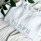 white crewneck sweatshirt with easter religious icons embroidered in silver sage green thread across the chest
