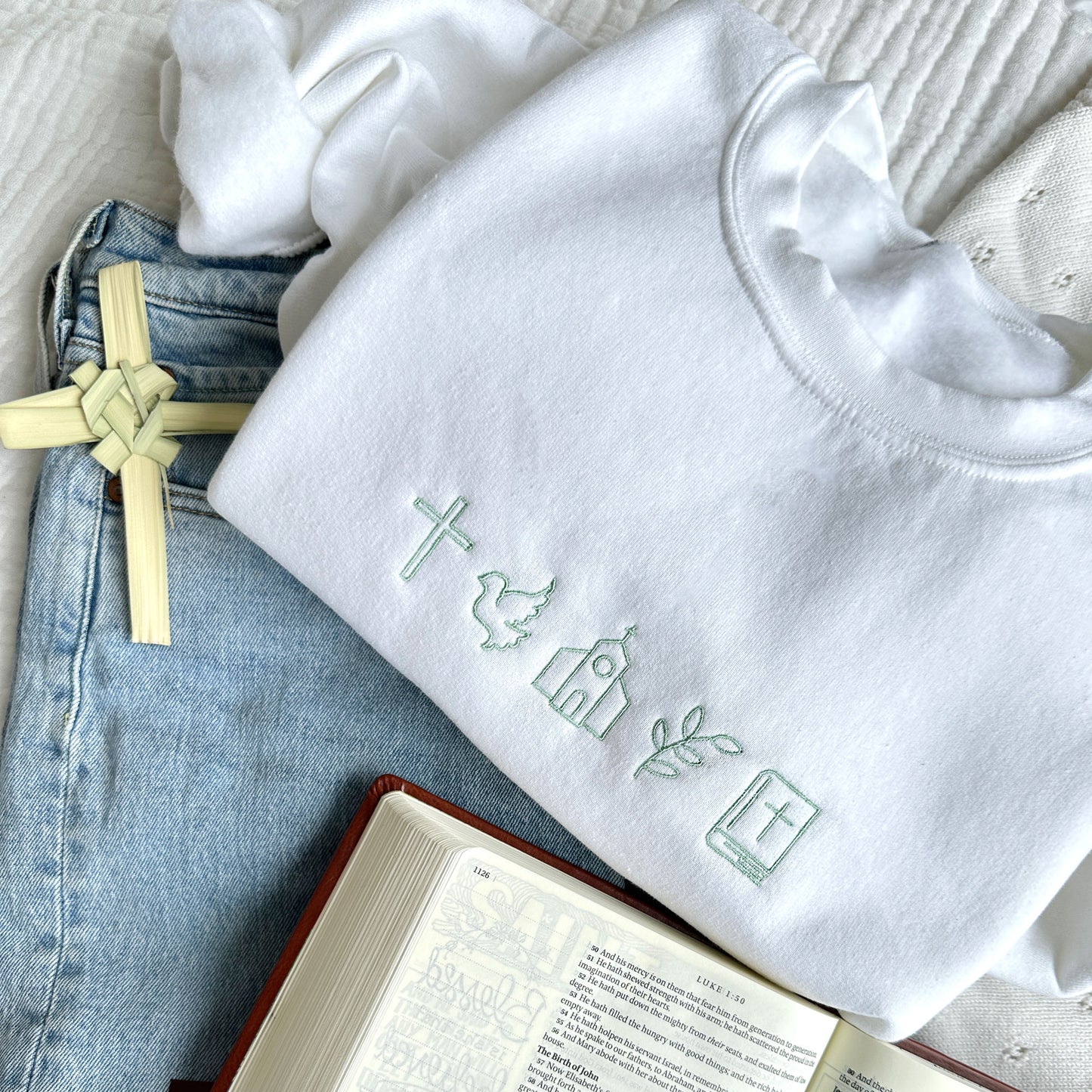 flat lay with jeans, a bible, palm branch, and a white crewneck sweatshirt. the sweatshirt has religious icons embroidered across the chest in silver sage thread