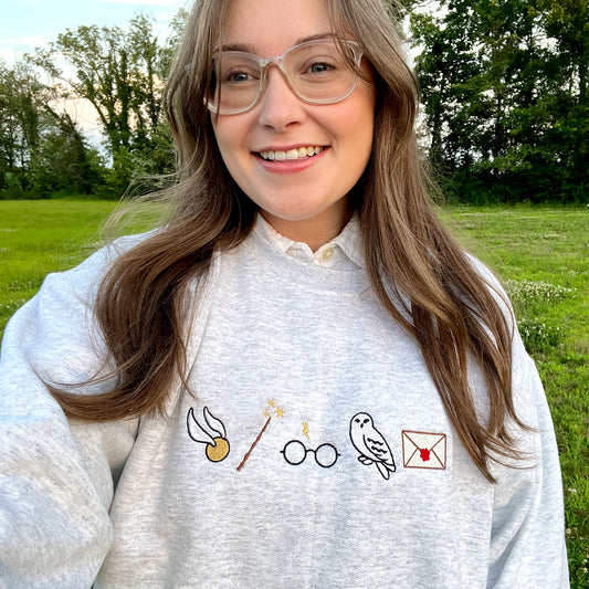girl wearing an ash gray sweatshirt with embroidered harry potter icons -- the golden snitch, wand, harry's glasses, hedwig, and an envelope 