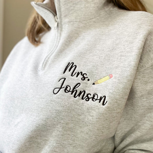 teacher wearing an ash gray quarter zip with a custom name embroidered design featuring a cute mini pencil icon embroidered next to it on the left chest