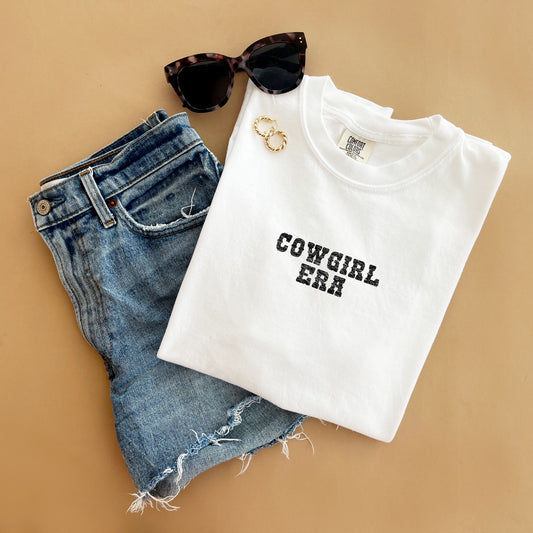 outfit layout featuring jean shorts, sunglasses, and a white tee with a distressed cowgirl era printed design