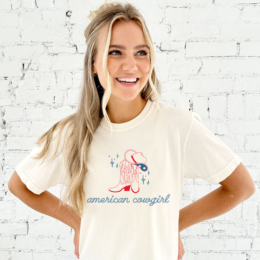 girl wearing an oversized ivory comfort colors t-shirt with a cute american cowgirl print in a rope font with cowgirl boots and a cowgirl hat