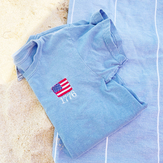 flat lay on a beach featuring a youth washed denim tee with a mini flag 1776 embroidered design on the left chest area
