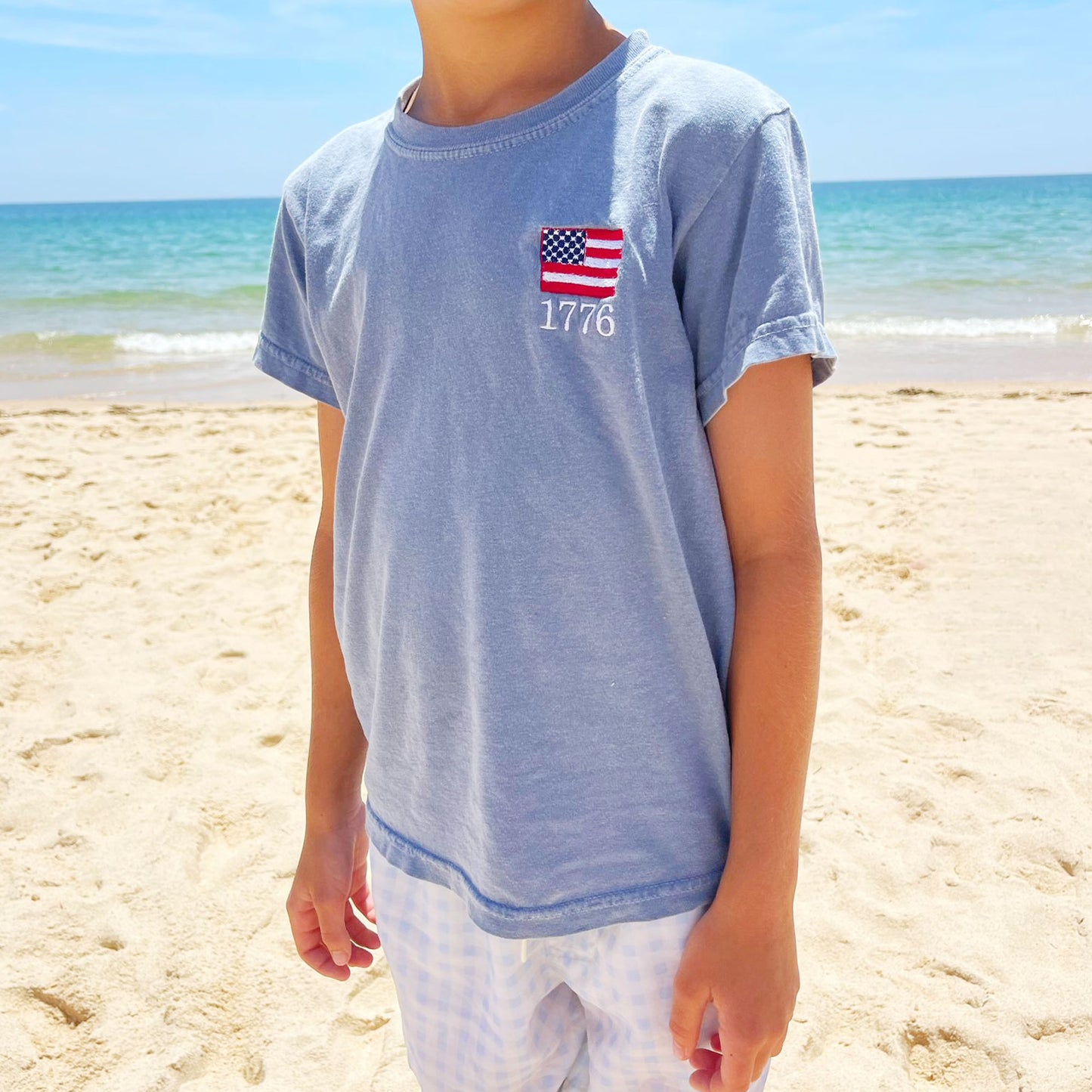 little boy wearing a washed denim comfort colors tee with a 1776 mini flag embroidery on the left chest standing on the beach