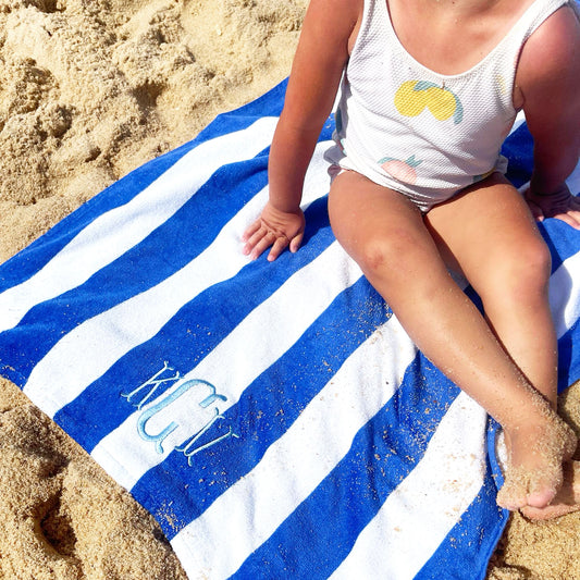 toddler girl sitting on a beach on top of a royal blue and white stripped beach towel with KCV monogram embroidered in powder Blue thread