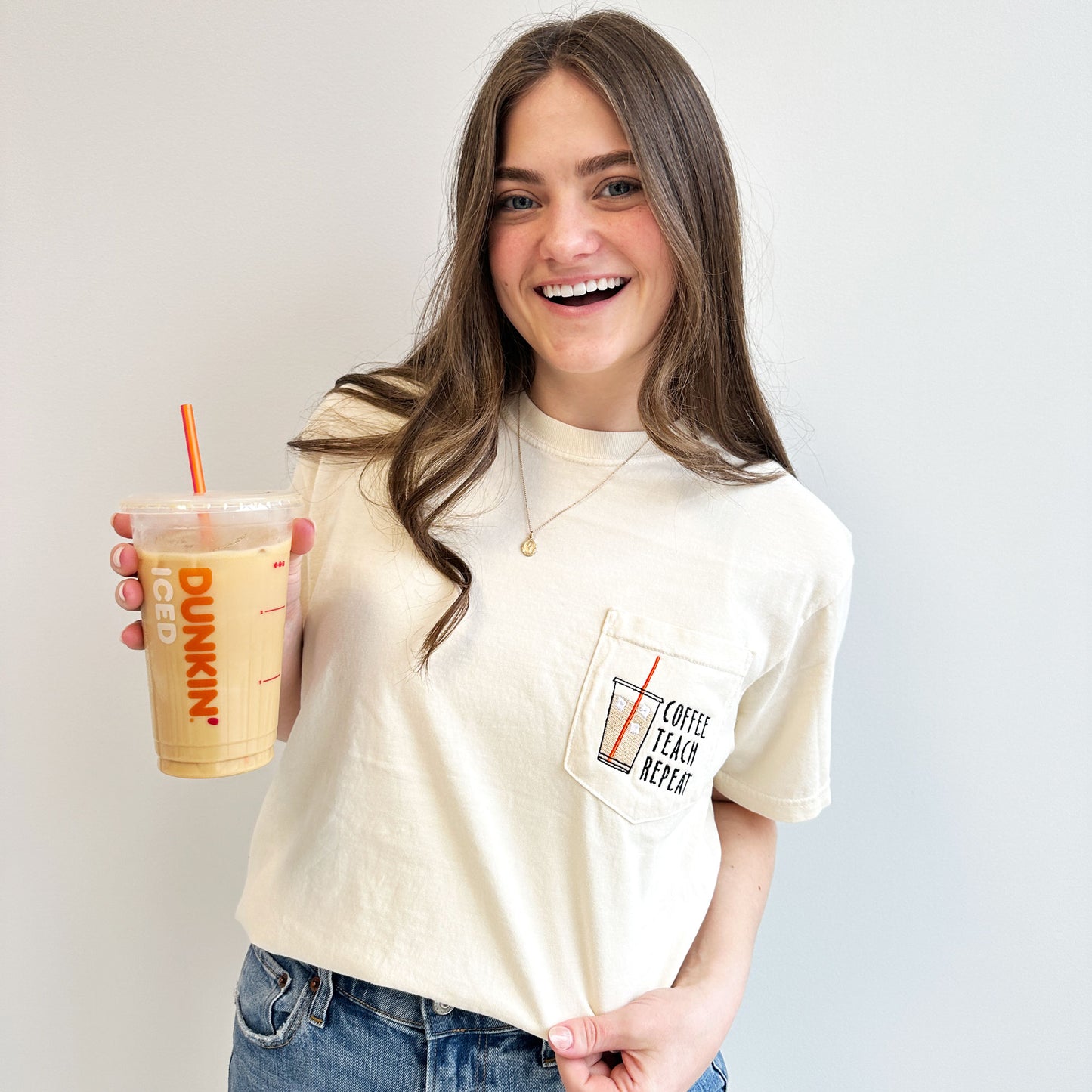 young woman wearing an ivory comfort colors pocket t-shirt with iced coffee and coffee teach repeat embroidered design on the pocket holding a dunkin iced coffee