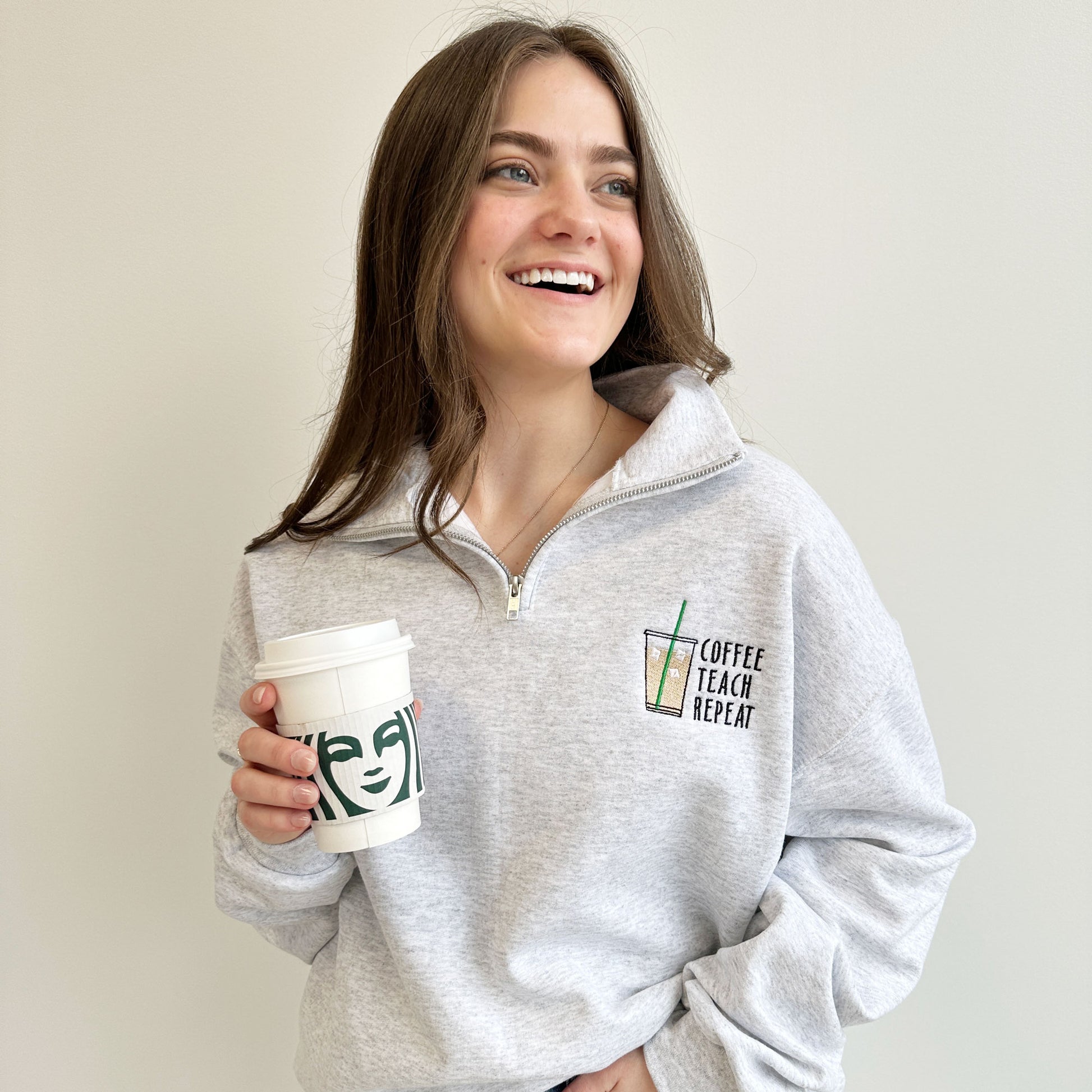 woman wearing a grey quarter zip sweatshirt with a coffee teach repeat embroidered design on the left chest