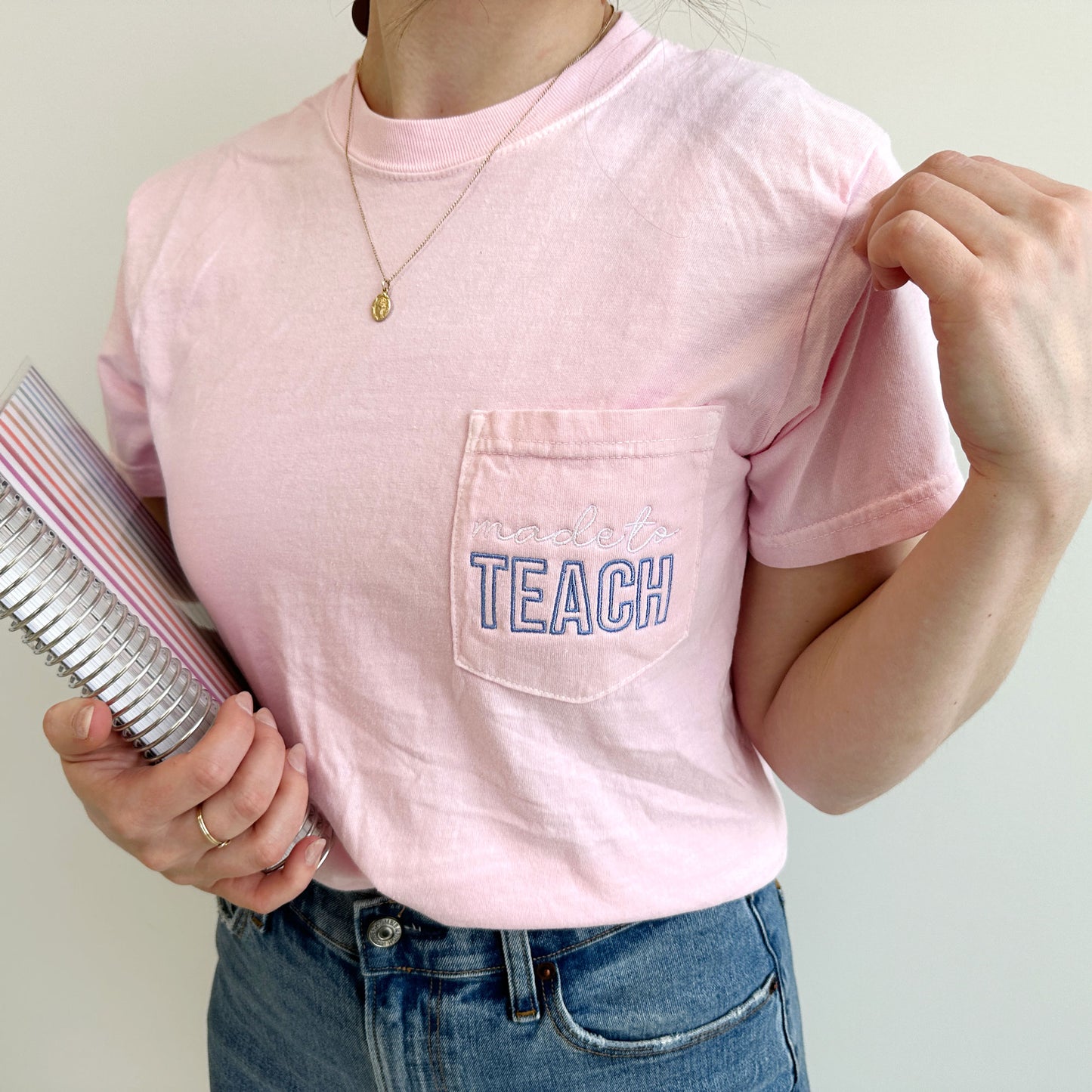 young woman wearing a blossom comfort colors t-shirt with made to teach embroidered design on the pocket in white and periwinkle threads