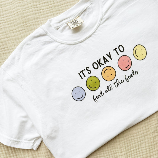 white crewneck t-shirt with a cute smiley face it's okay to feel all the feels printed design