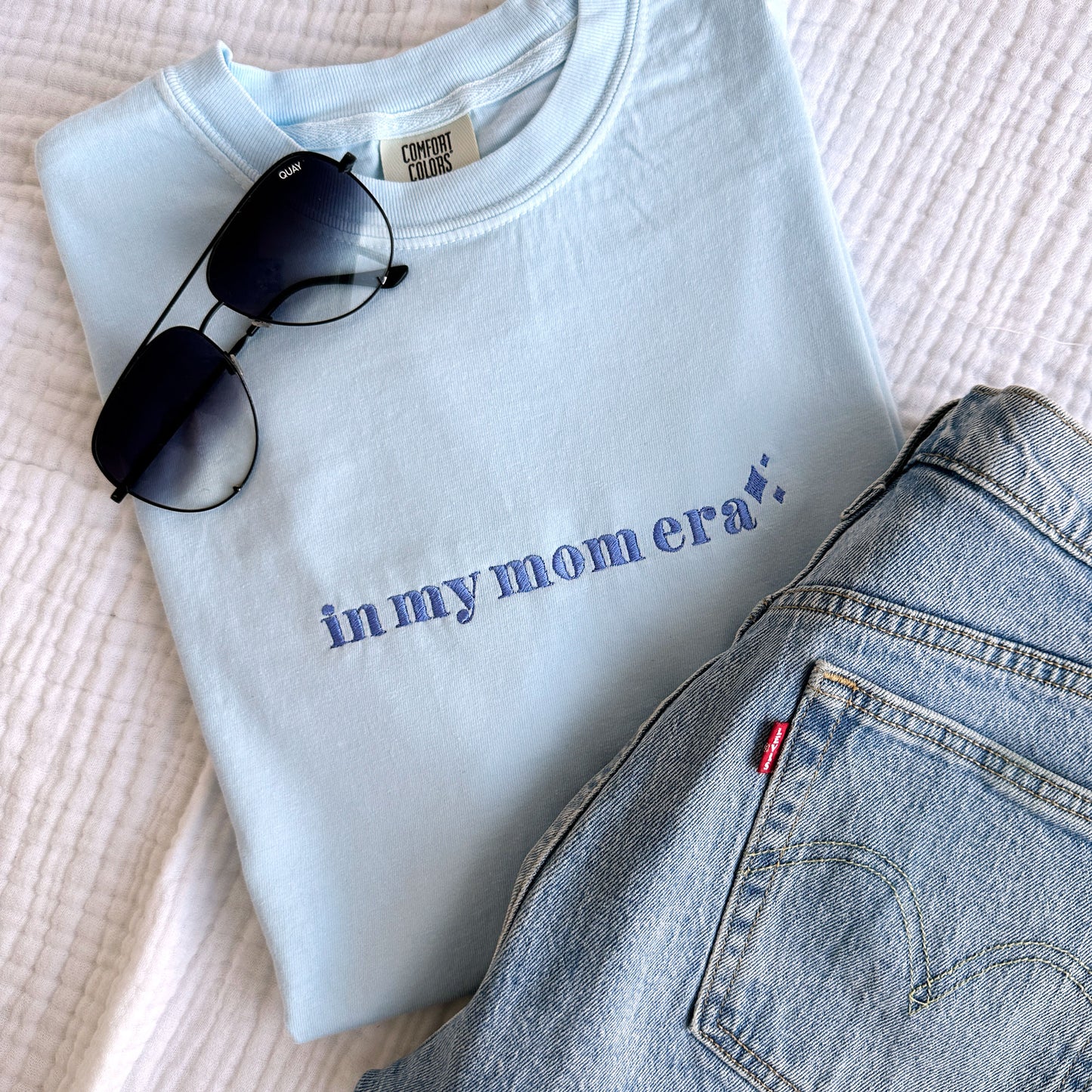Chambray comfort colors t-shirt with embroidered in my mom era design across the chest in periwinkle thread styled with jeans and sunglasses