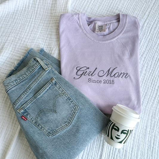 flat lay of an orchid comfort colors t-shirt with embroidered Girl Mom since custom date design across the chest in smoky orchid thread styled with jeans and a Starbucks cup