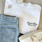 styled flat lay of a white comfort colors tshirt with embroidered stay at home moms club design small on the left chest in hunter green thread with jeans  and sneakers