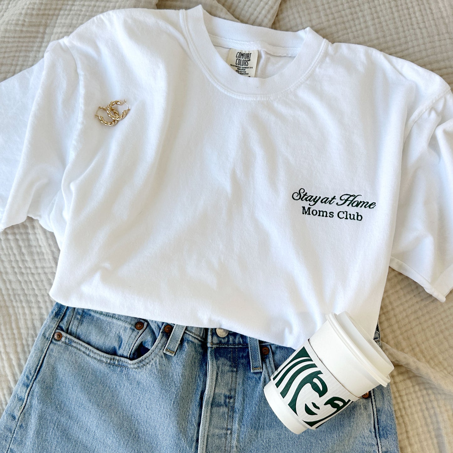 styled flat lay of a white comfort colors tshirt with embroidered stay at home moms club design small on the left chest in hunter green thread with jeans  and a starbucks cup